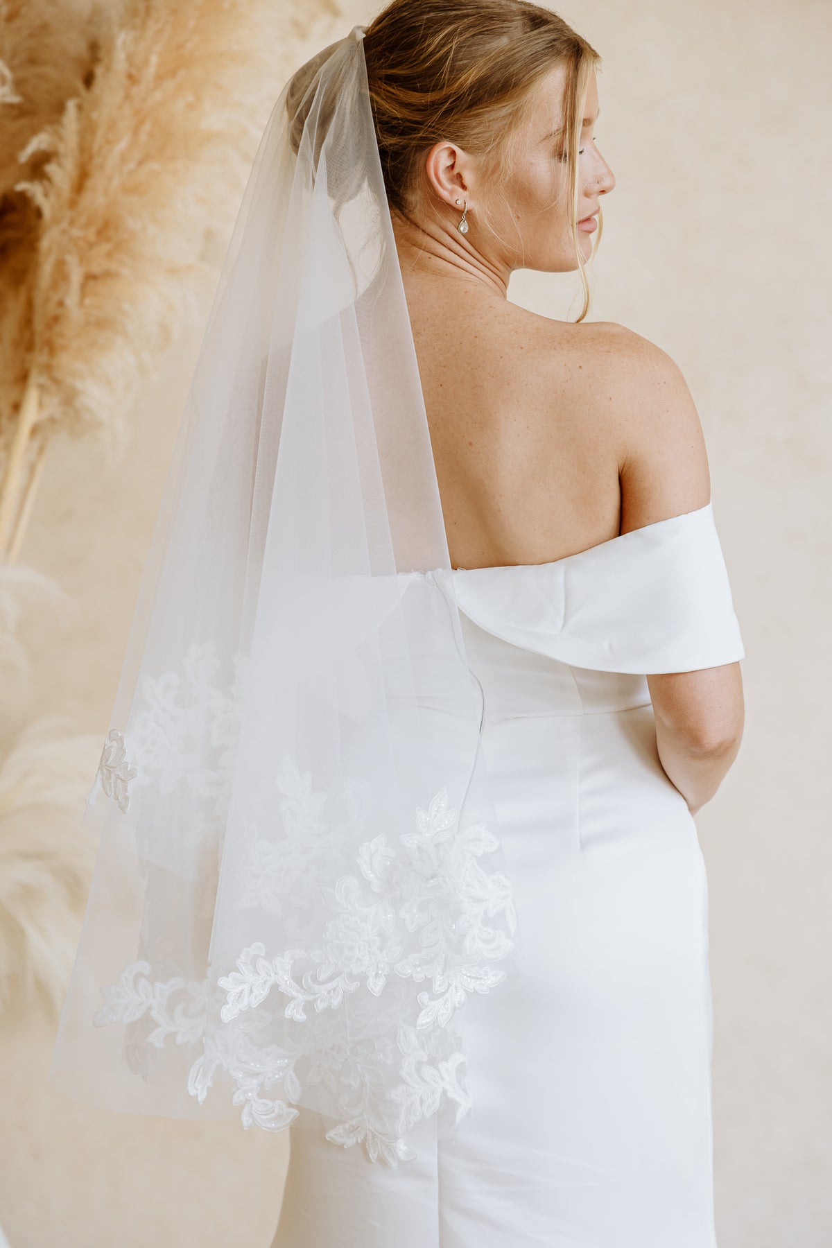 A bride shown from the back wearing a white off-the-shoulder wedding dress and a long sheer tulle veil with floral lace trim, posed against a warm beige background with pampas grass.