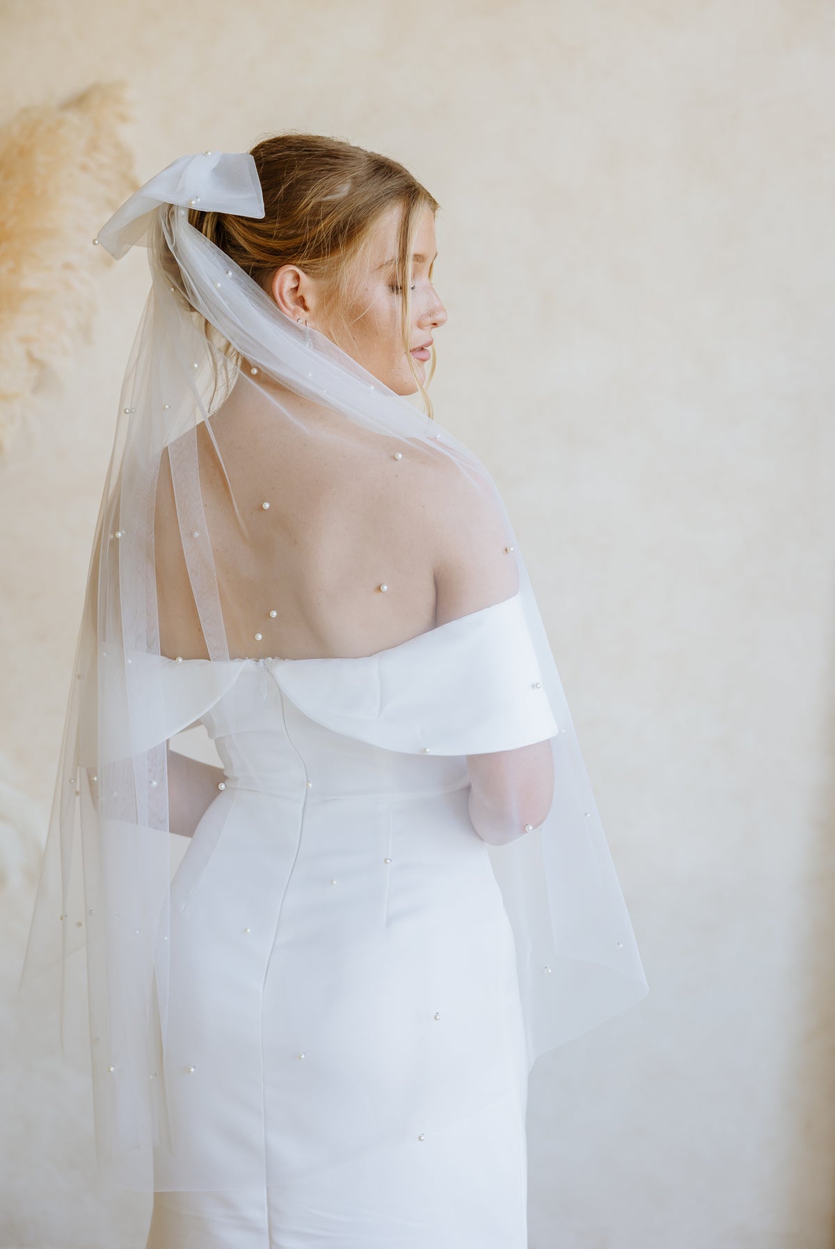 A bride in a white off-the-shoulder wedding dress with a sheer embellished veil, shown from the back against a soft beige wall.