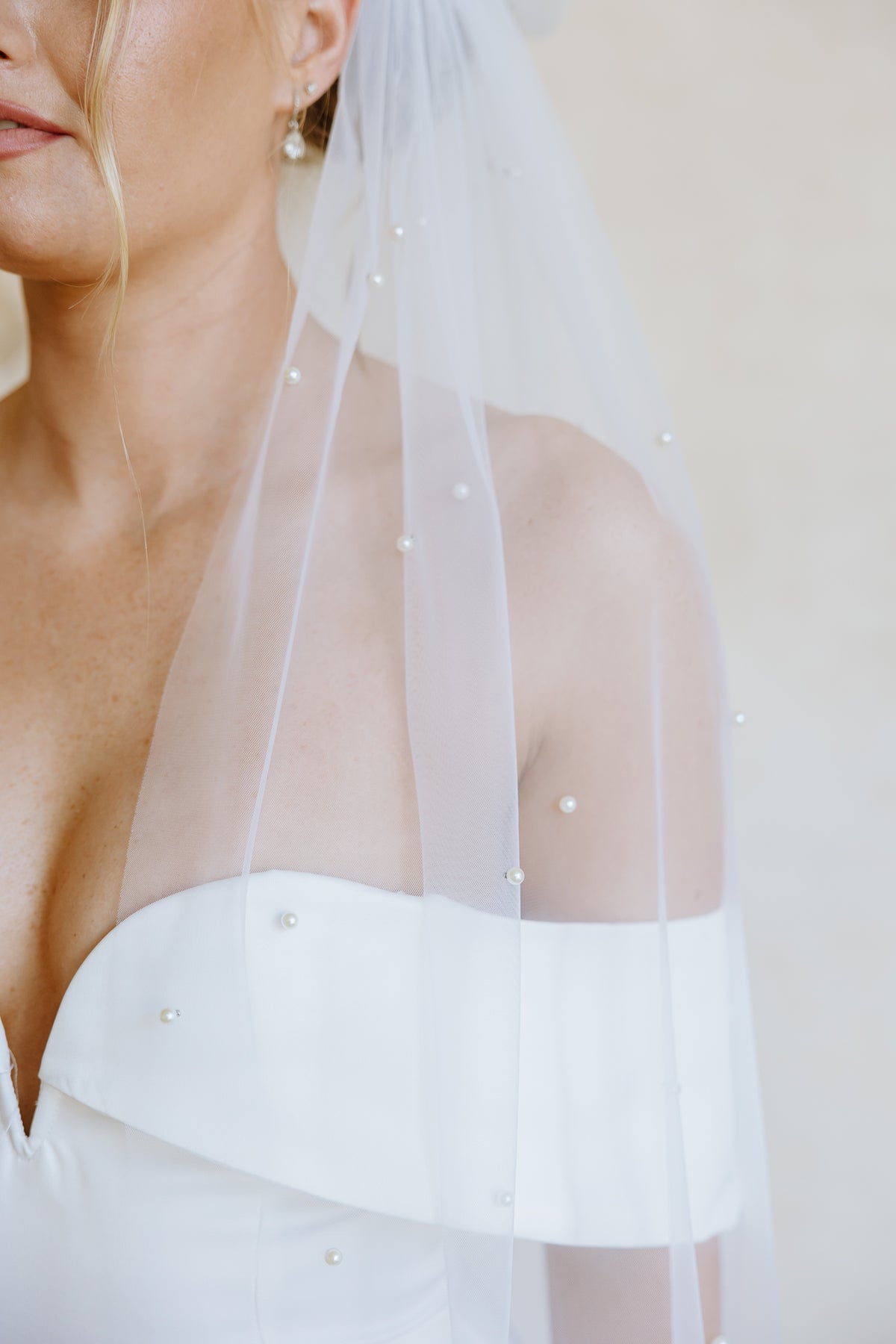 A close-up of a bride wearing a sheer white tulle veil adorned with small pearl-like beads, paired with a white strapless wedding dress against a soft beige background.