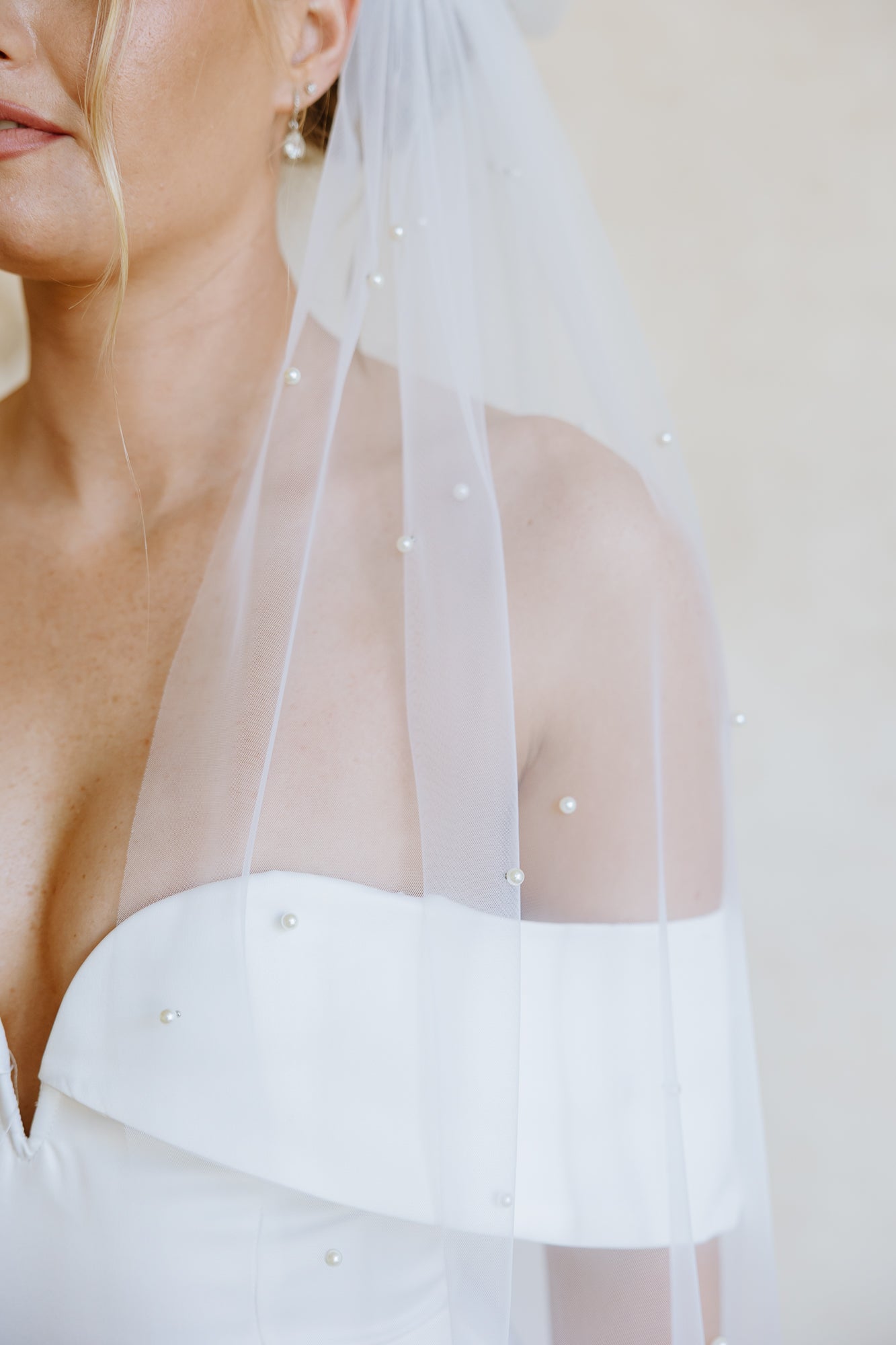 A bride in a white off-the-shoulder wedding dress with a sheer embellished veil, shown from the back against a soft beige wall.