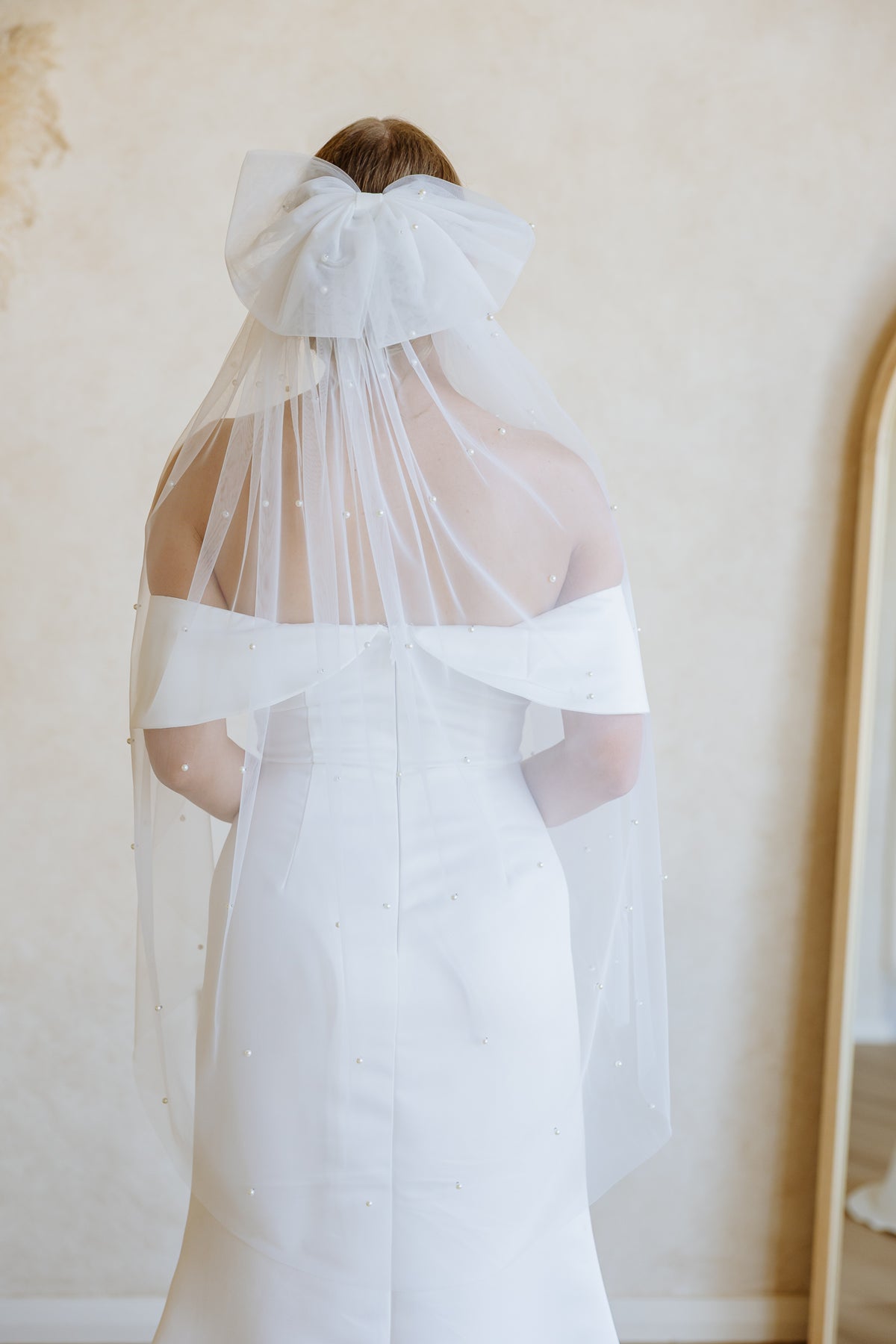 A bride viewed from behind wearing a white wedding dress and a long sheer veil with a large bow at the top, standing indoors against a warm beige wall.