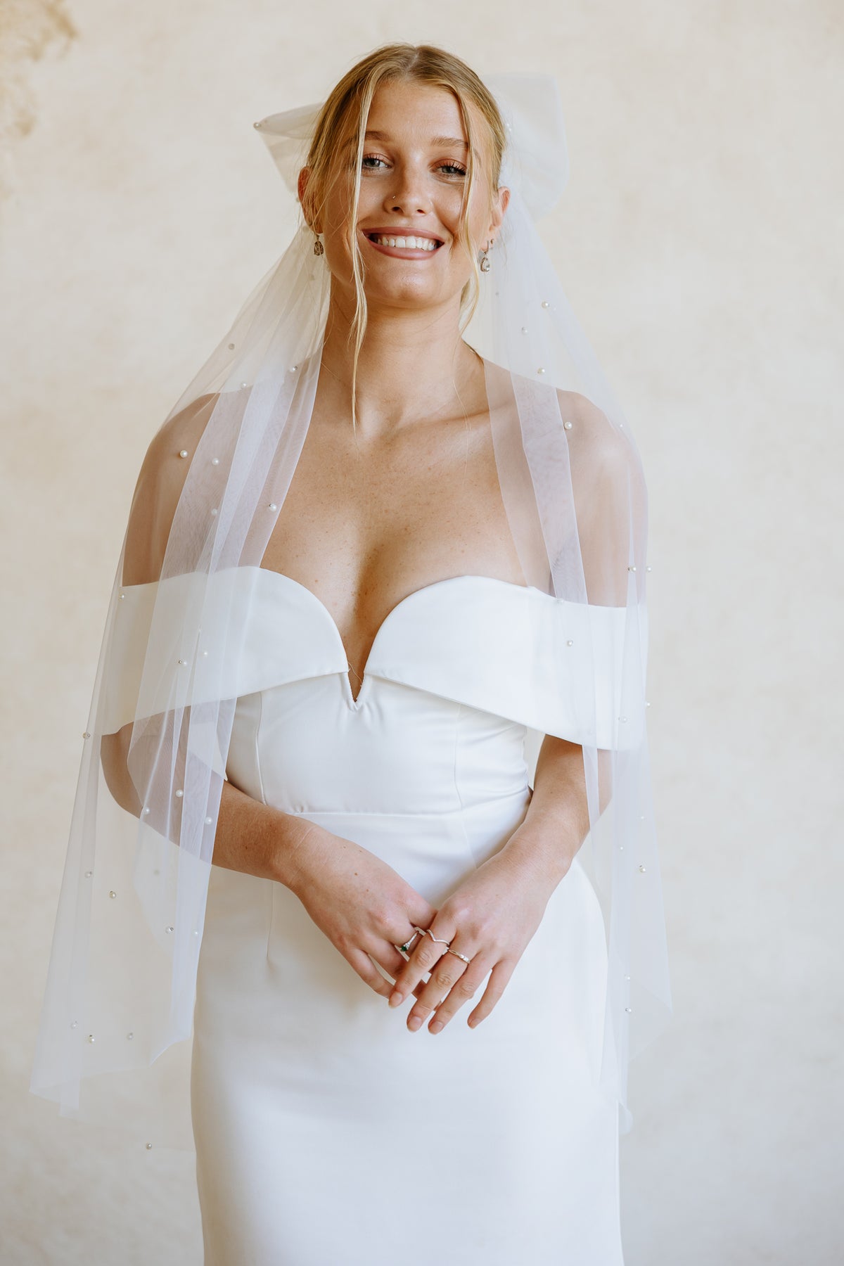 A smiling bride wearing a white strapless wedding dress with a sheer white veil, standing against a warm beige textured wall.