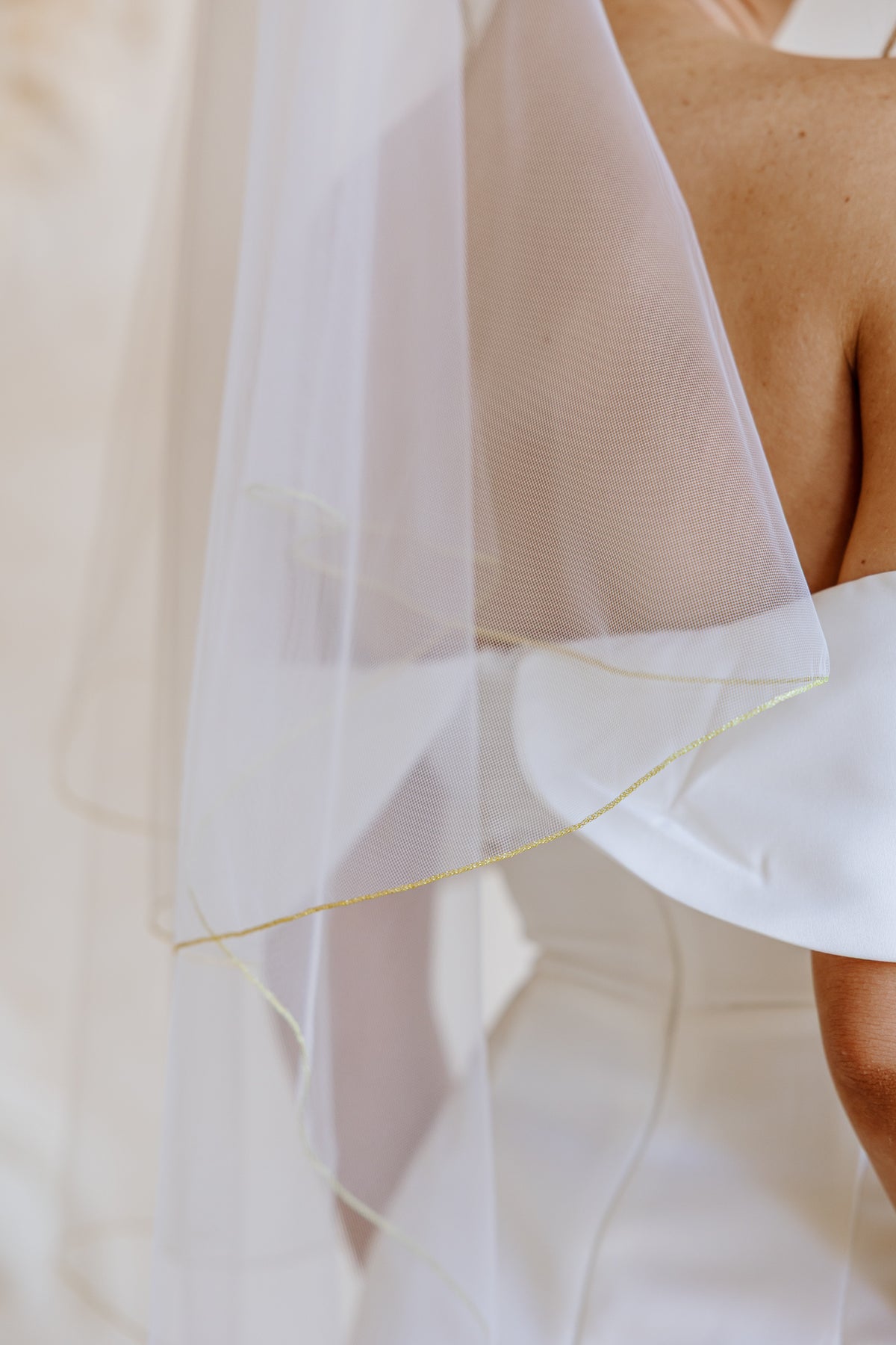 Close-up of a white bridal veil with a golden trim draped over a bride's shoulder, with part of her bare back and white wedding dress visible against a soft cream background.
