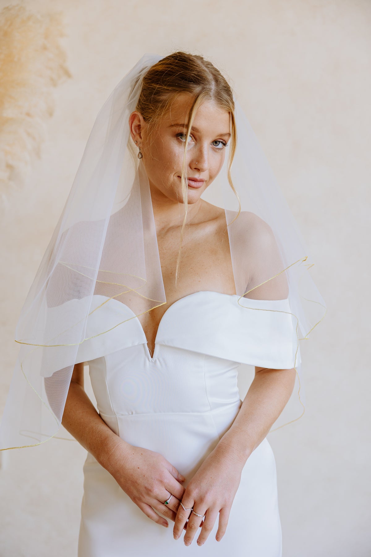 A woman wearing a white off-the-shoulder wedding dress and a sheer white veil with gold trim detail, standing against a warm beige textured wall.