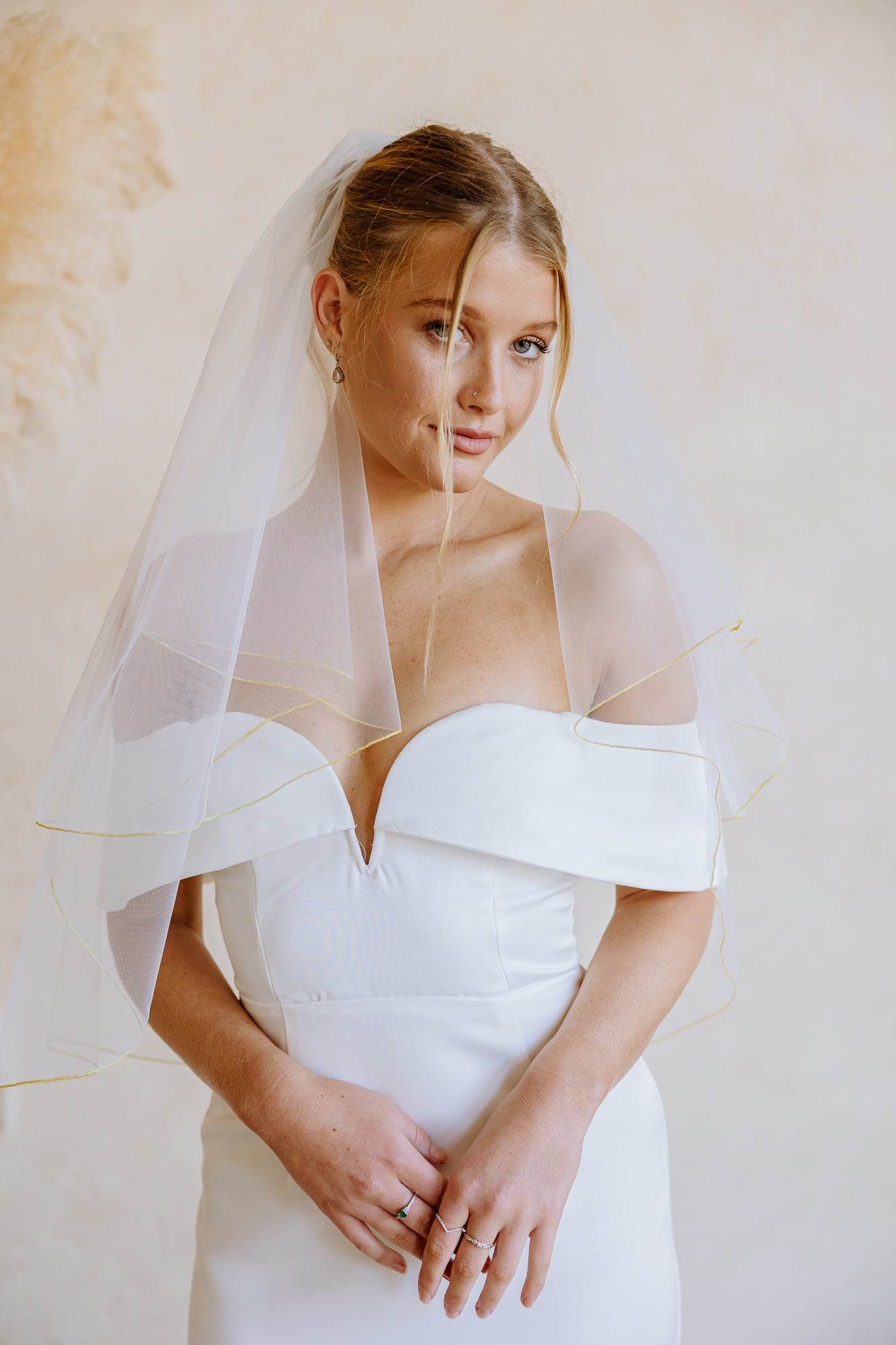 Close-up of a white bridal veil with a golden trim draped over a bride's shoulder, with part of her bare back and white wedding dress visible against a soft cream background.