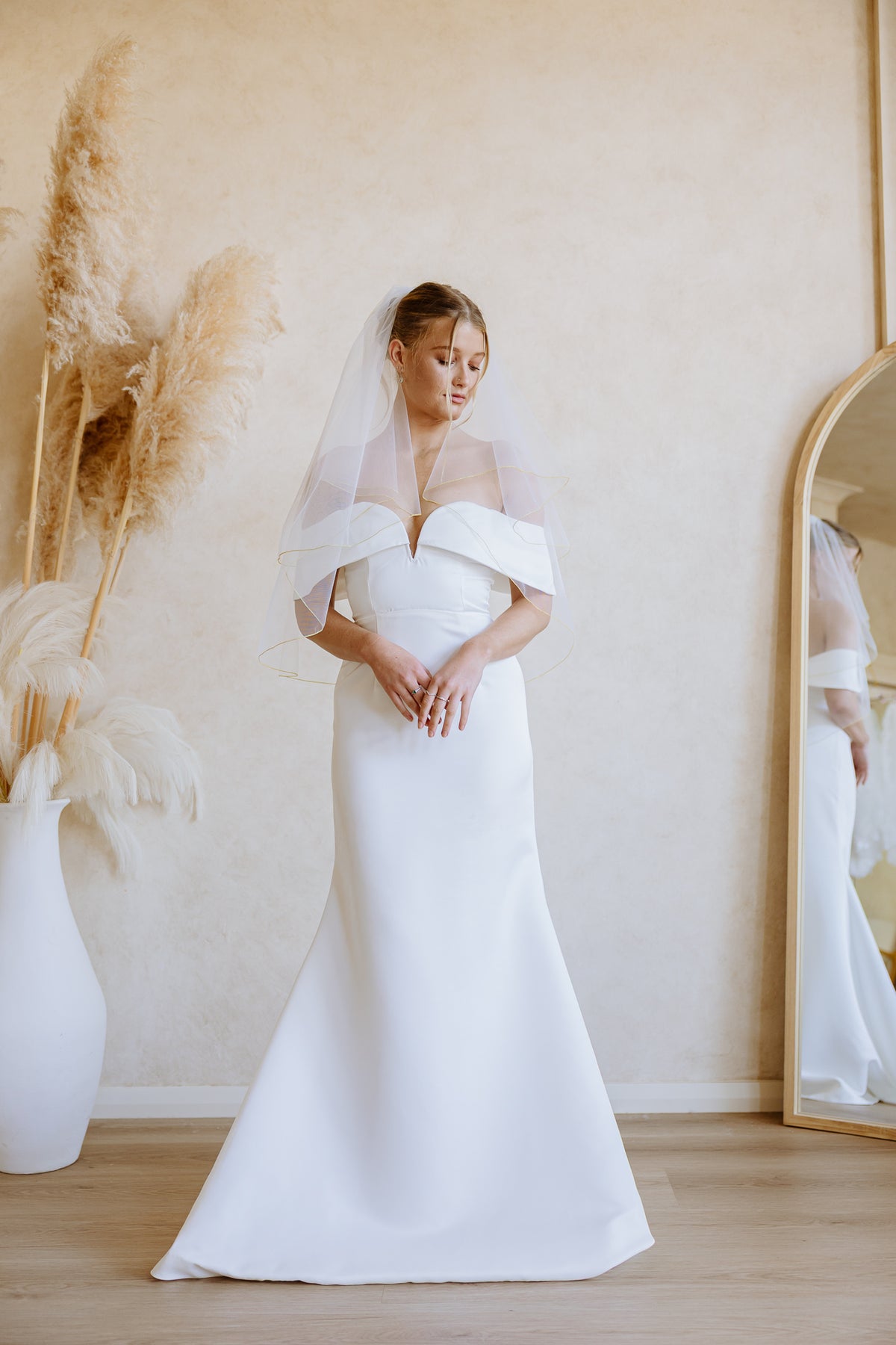 A woman wearing a white off-the-shoulder mermaid-style wedding dress with a sheer veil stands in a softly lit room with a beige wall, wood floor, pampas grass in a white vase, and a mirror showing part of her reflection.