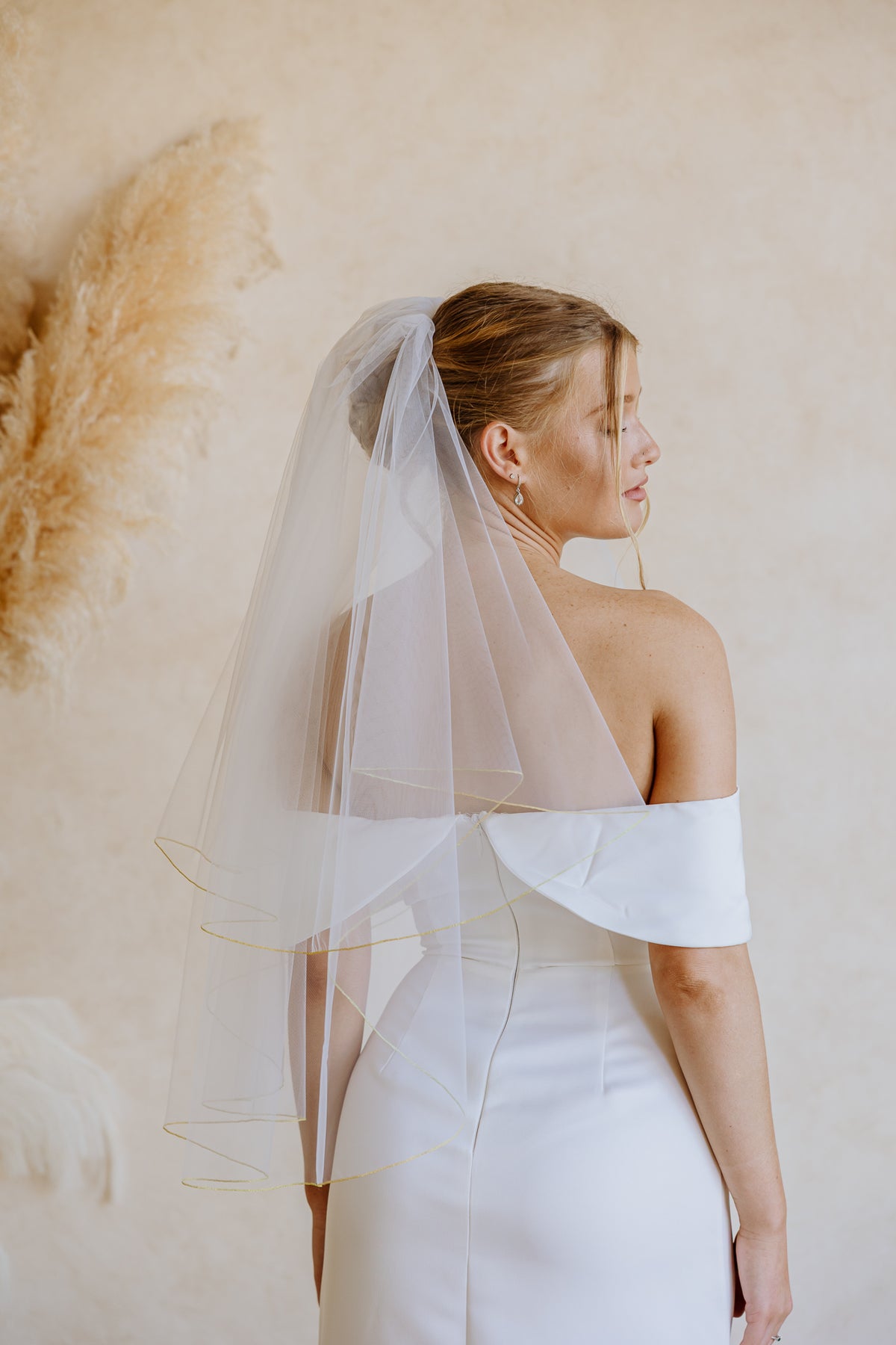 A woman in a white off-the-shoulder wedding dress and long sheer veil, shown from the back in profile against a warm beige wall with pampas grass decor.