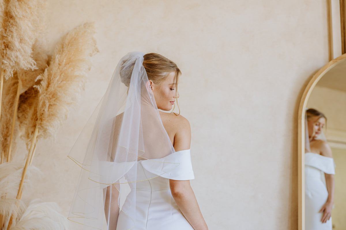 A bride in a white off-the-shoulder wedding dress with a long sheer veil with a golden trim, shown from the back in a softly lit room with a mirror and pampas grass decor.