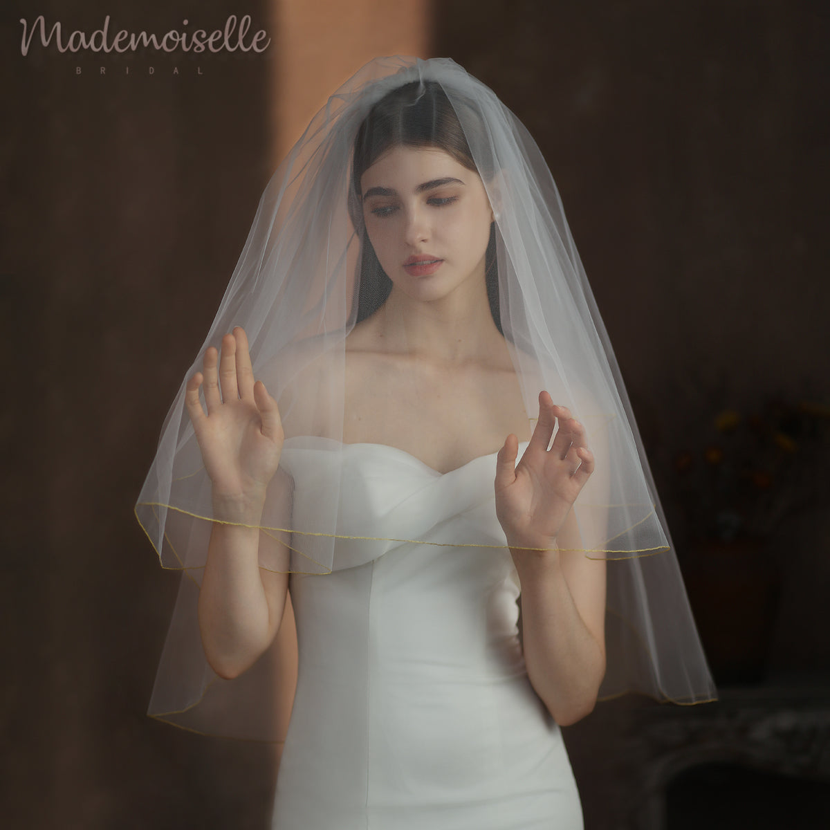 A bride in an exquisite white wedding veil featuring golden threaded trim, gently clasping her hands in a serene and elegant pose.