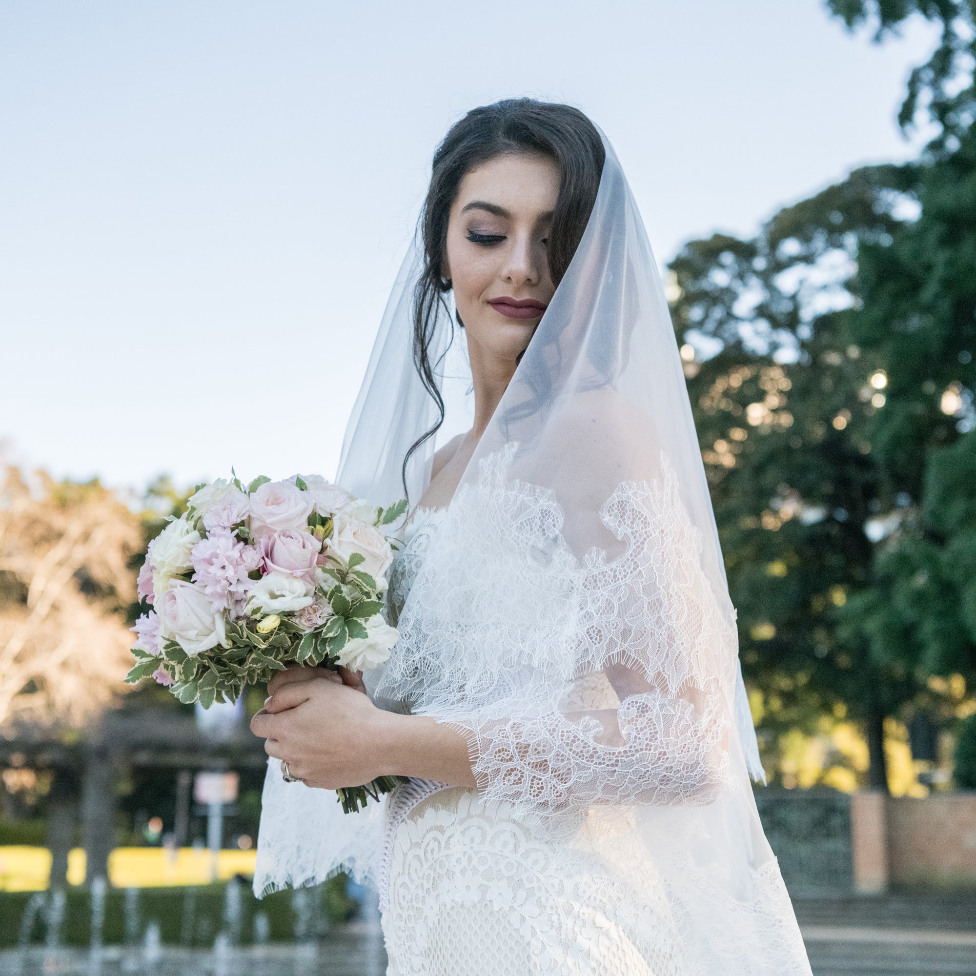 A bride elegantly poses in front of a beautiful fountain, surrounded by lush greenery in a serene park setting.