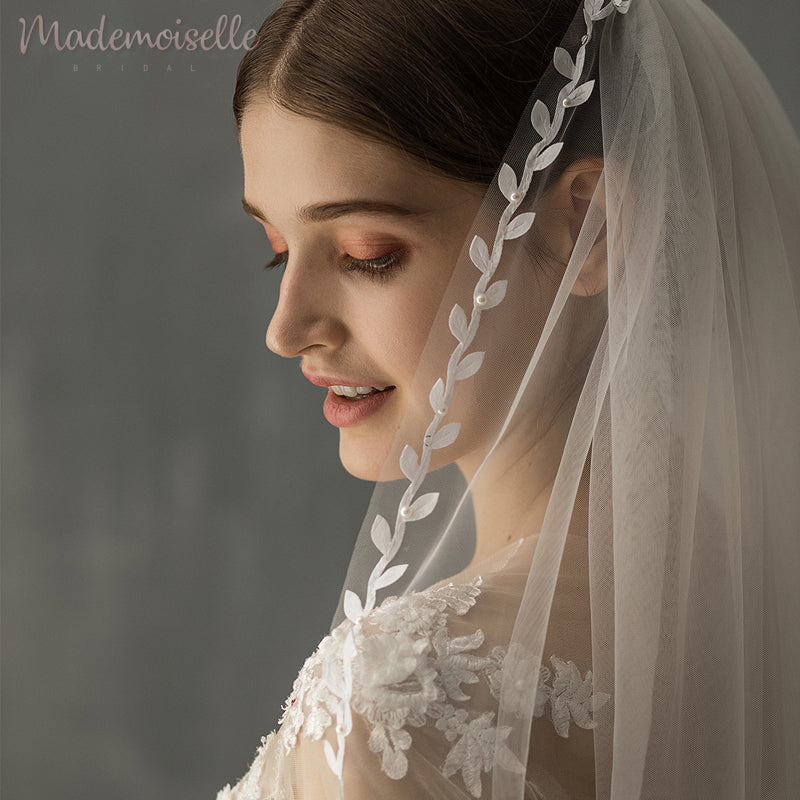 Bride in a lacy white dress and veil stands with her back to the camera against a gray background.